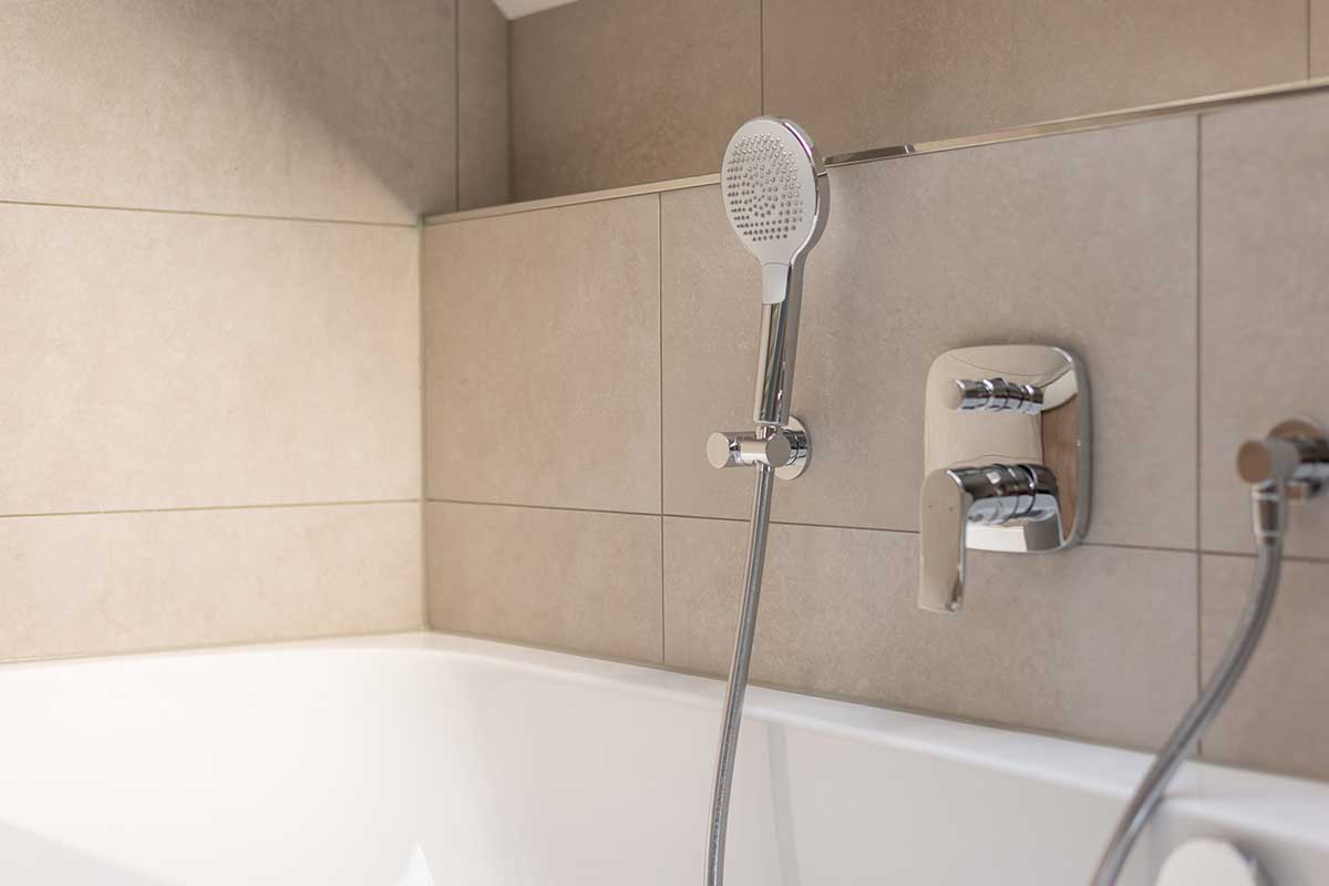 Moderne Badewannenarmatur mit Handbrause vor beigefarbenen Fliesen.