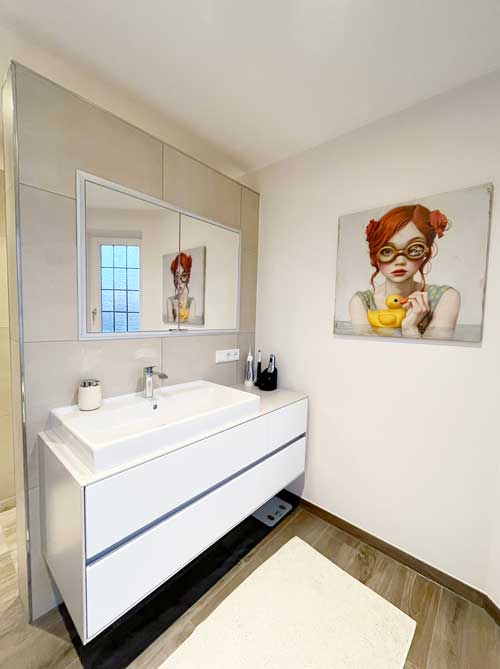 Modernes Badezimmer mit weißem Waschtisch und großer Spiegelfläche, kombiniert mit warmen Wand- und Bodentönen. An der Wand hängt ein humorvolles Porträt eines Mädchens mit rotem Haar und Quietscheente – ein stilvoller Blickfang.