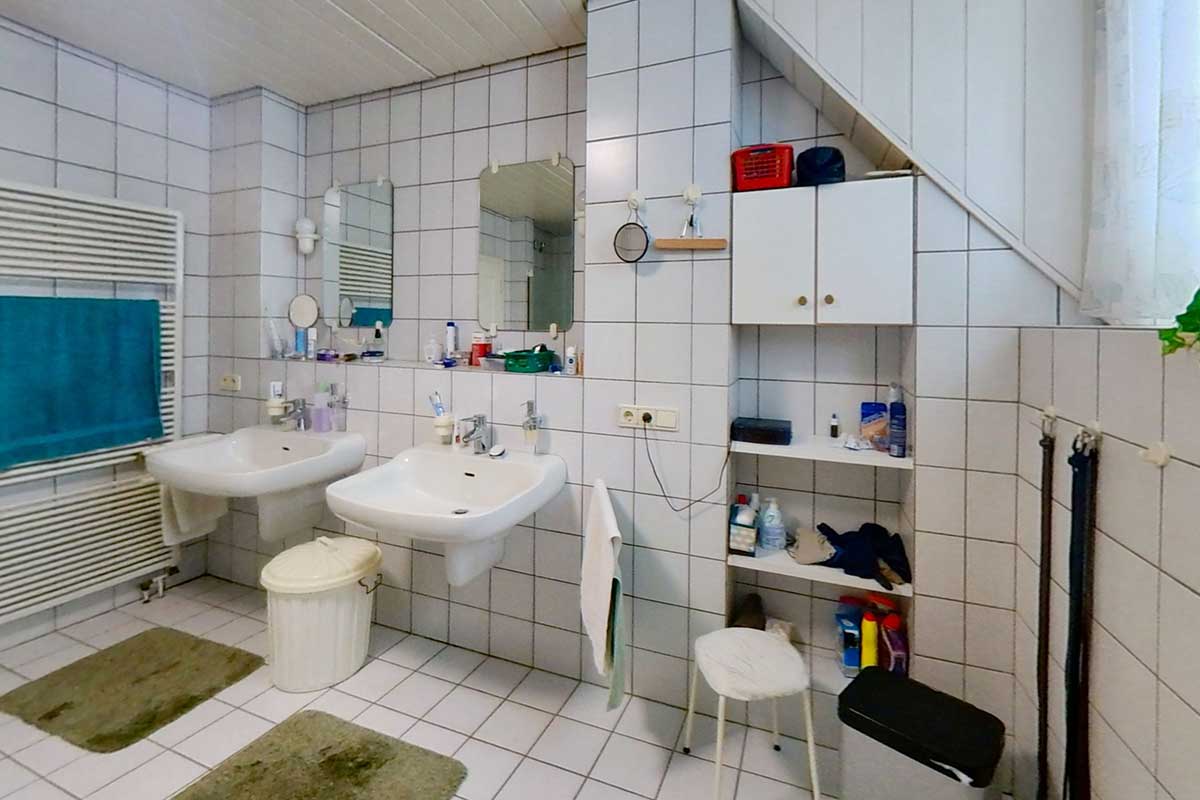 Altes Badezimmer mit weißen Fliesen, zwei Waschbecken, einfacher Möblierung und sichtbarer Dachschräge