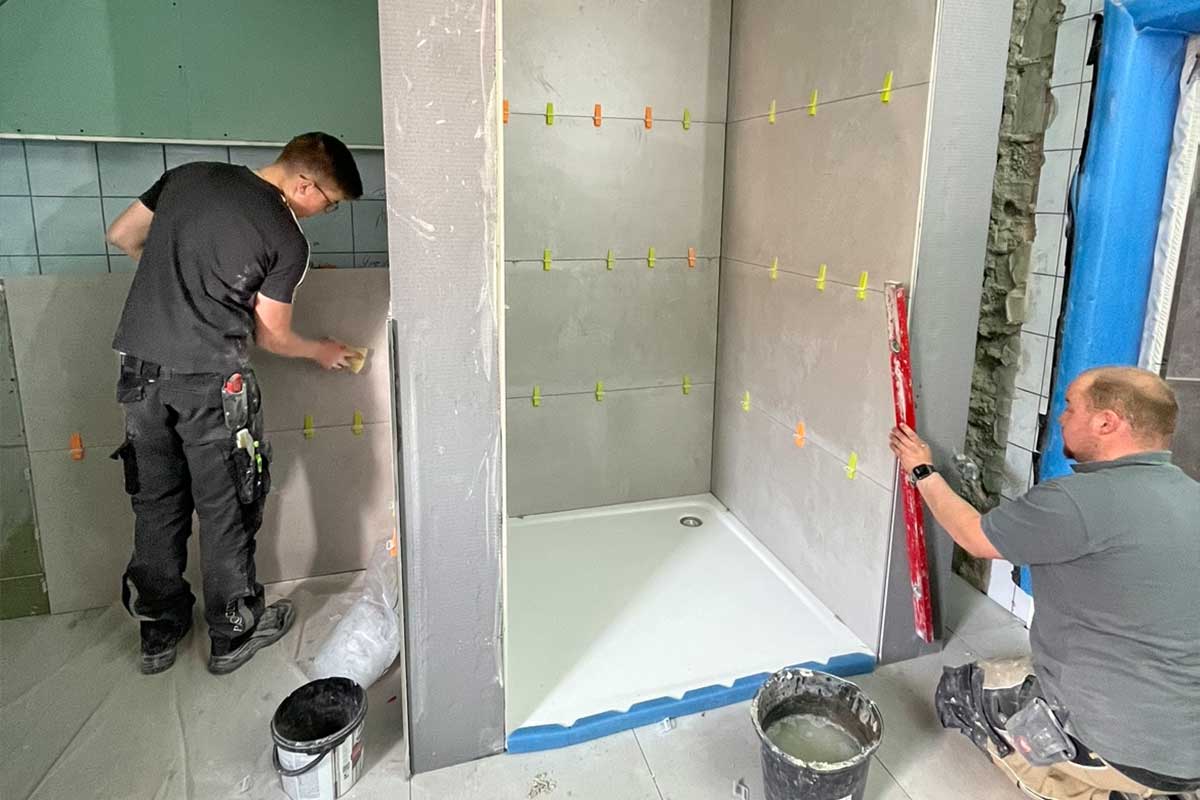 Fliesenarbeiten im Gange: Zwei Handwerker beim Verlegen großformatiger Wandfliesen in der zukünftigen Dusche.