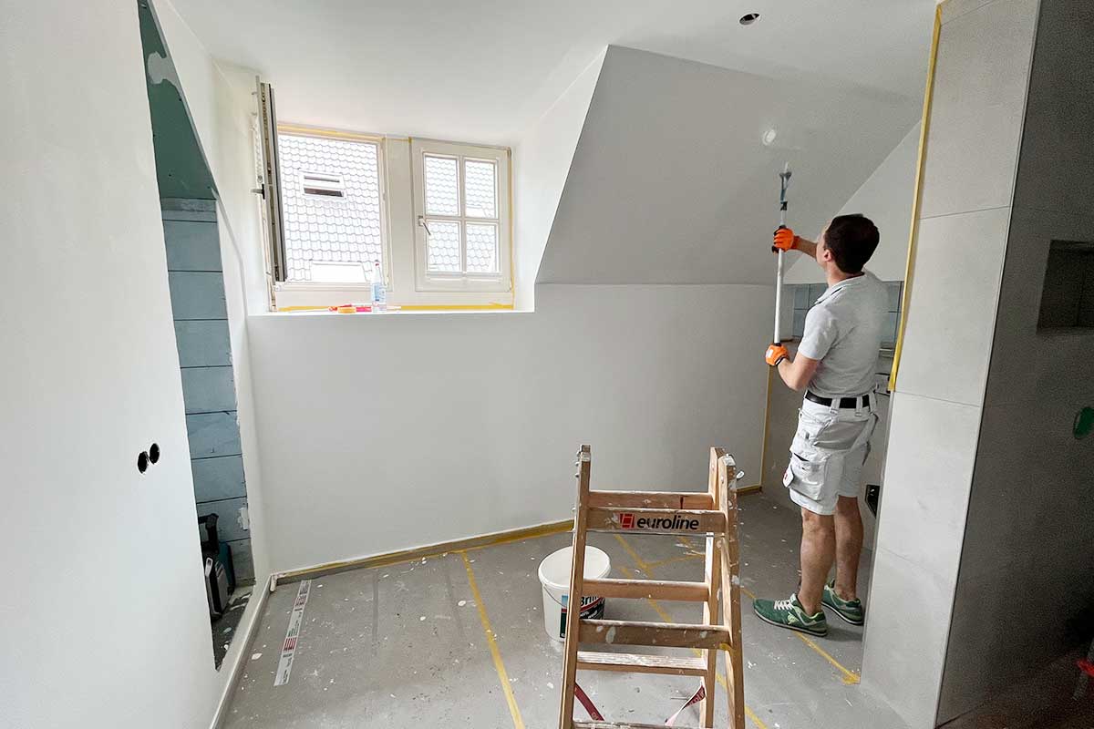 Malerarbeiten im fast fertigen Badezimmer – Wände werden weiß gestrichen, Fensterbereich ist abgeklebt.