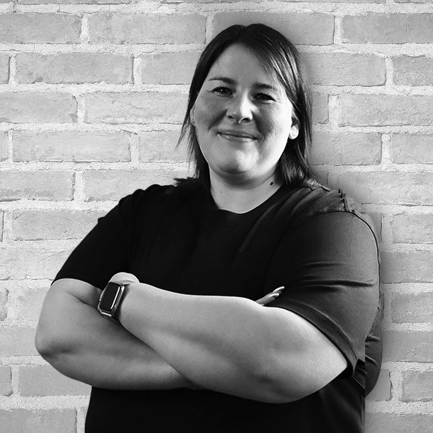 Porträt von Jana Panten, Kauffrau für Büromanagement, vor einer hellen Backsteinwand, mit verschränkten Armen und freundlichem Lächeln.